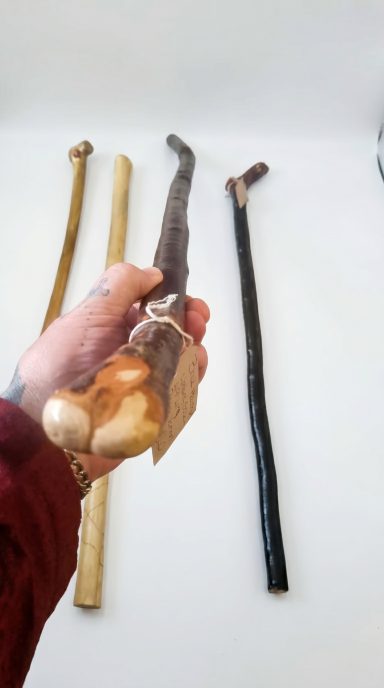Dark Brown Handcrafted Walking Stick Dark brown handcrafted wooden walking stick with maker’s tag and artisan staffs in the background